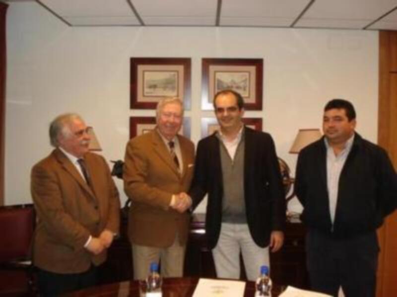 Cuatro hombres en una reunión formal, dos de ellos saludándose con un apretón de manos. La imagen muestra una oficina o sala de conferencias con decoración y mobiliario típicos.