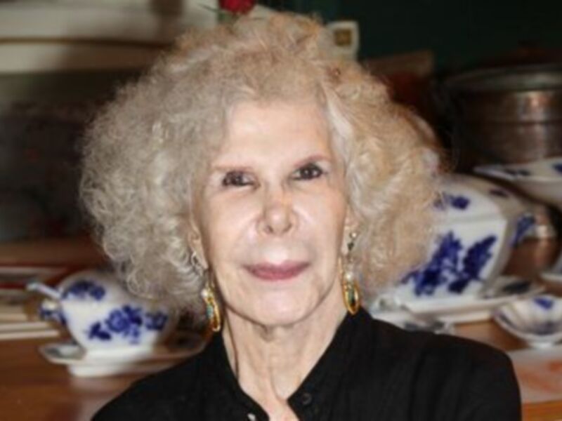 Una mujer con cabello blanco y una sonrisa amable, posando frente a un fondo de porcelana azul y blanca.