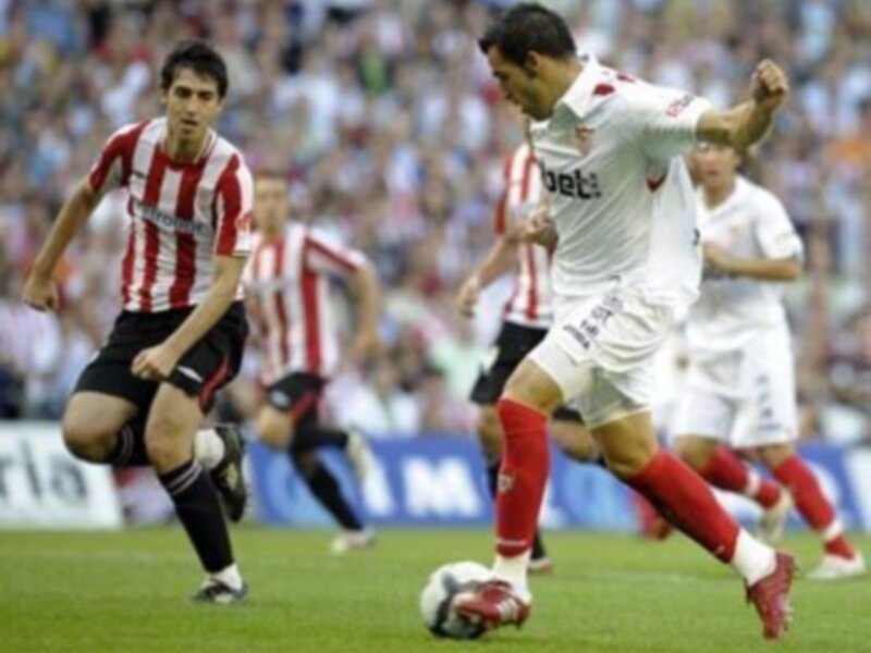 Un juego de fútbol en curso con jugadores en acción, uno de ellos con una camiseta roja y blanco y otro con una camiseta blanca y roja, ambos en un campo de césped verde.