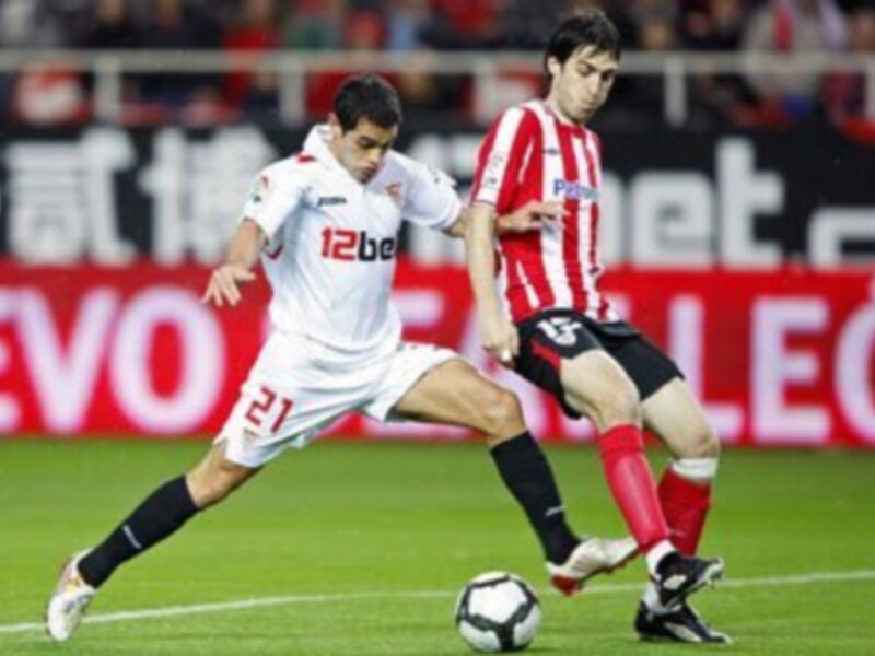 Dos jugadores de fútbol en acción durante un partido, uno con una camiseta blanca y otro con una camiseta roja y blanca. El balón está en el aire, indicando un momento de juego intenso.