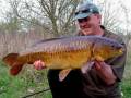 Un hombre sostiene con orgullo un gran pez carpa de color dorado brillante, posiblemente una especie de carpas de río. El fondo muestra un paisaje natural con árboles y vegetación, indicando que la imagen fue tomada en un entorno al aire libre.