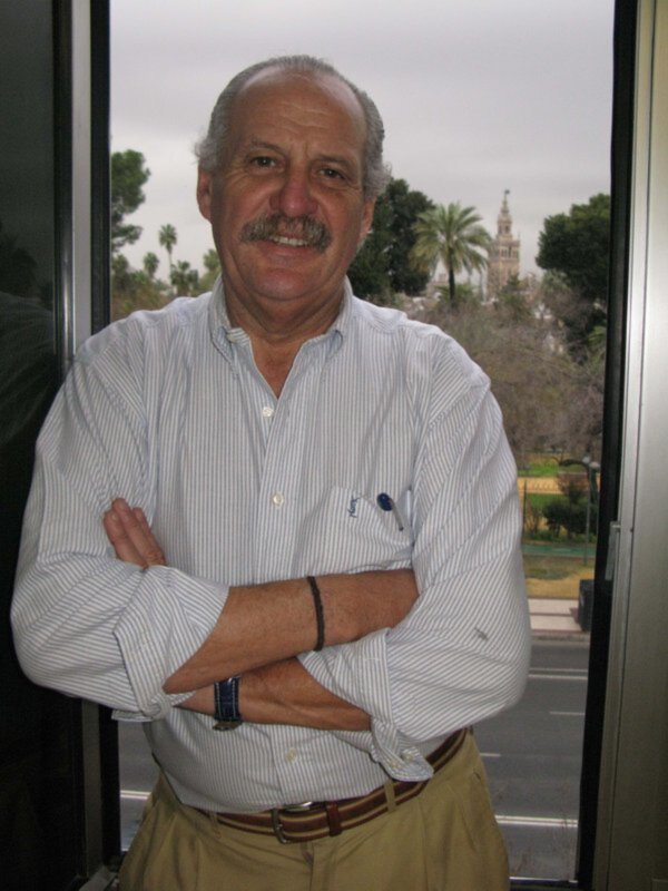 Un hombre con barba y bigote, vistiendo una camisa blanca con botones y pantalones amarillos, posa frente a un cristal que revela una vista de la ciudad con palmeras y un edificio histórico en el fondo.