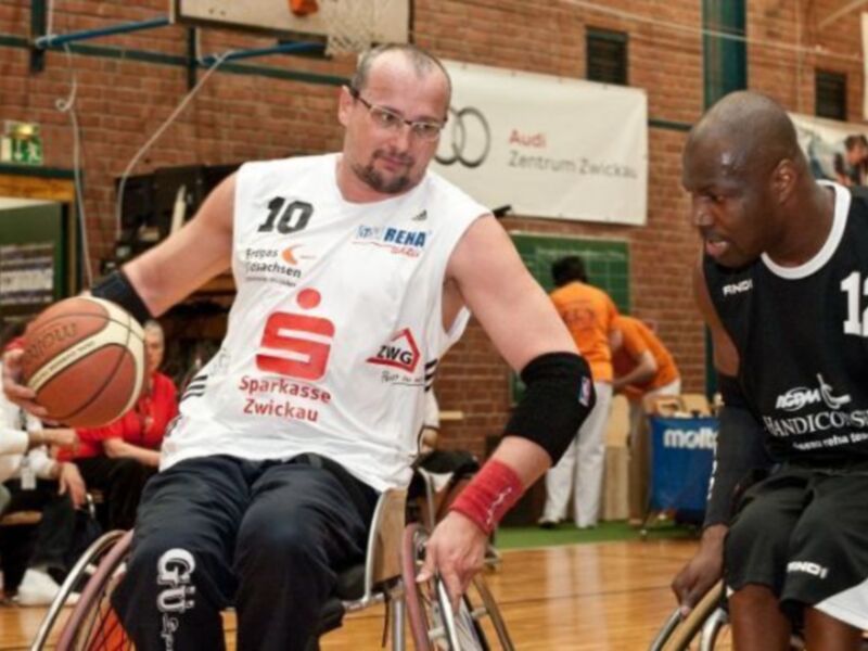 Un hombre en silla de ruedas juega baloncesto, con un equipo de uniforme blanco y rojo. Detrás de él, otro jugador en silla de ruedas con una camiseta negra y el número 12. En el fondo, un banner de Audi y una puerta con una pancarta.