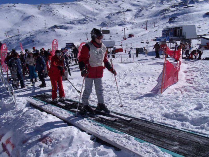 Un esquiador en rojo con una chaqueta blanca y pantalones azules, camina sobre un camino de esquí con otros esquiadores en el fondo. La imagen muestra una estación de esquí con varios esquiadores y un telecabina en el horizonte.