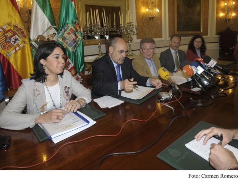 Una reunión formal en un salón con banderas españolas y personajes sentados alrededor de una mesa, rodeados por micrófonos.