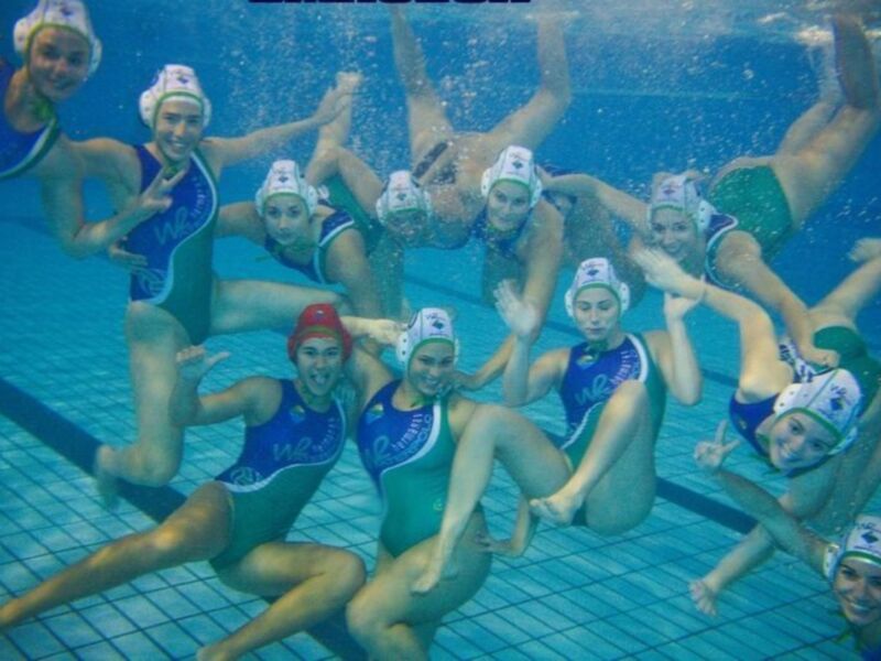 Una formación de waterpolo femenino bajo el agua, con jugadoras en trajes azules y verdes, acuáticas y sincronizadas.