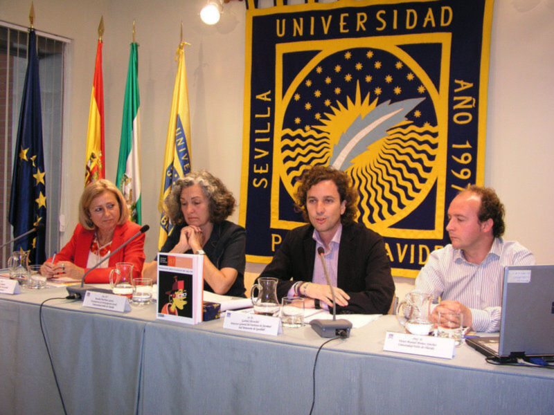 Una conferencia de prensa en la Universidad de Sevilla, con tres personas sentadas en una mesa, frente a una bandera de la universidad y banderas de España y Andalucía.