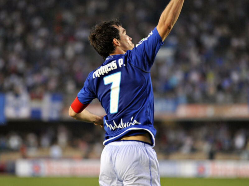Un jugador de fútbol levanta la camiseta azul con el número 7 y el nombre 'Francis P.' en la parte superior, mientras el público se desvanece en el fondo.
