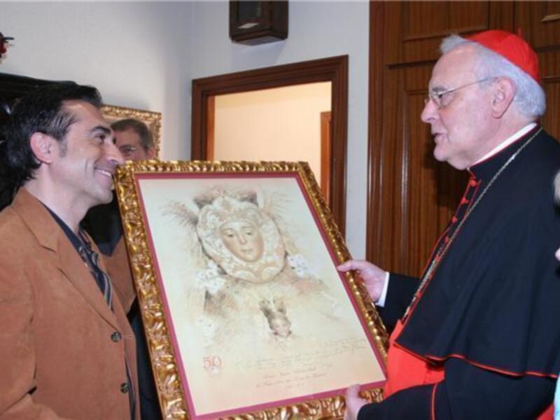 Un hombre en traje marrón sostiene una pintura que muestra a un personaje con sombrero y tocado, mientras otro hombre en traje negro y capa de cardenal la mira. La escena parece ser una reunión o presentación formal, posiblemente en un evento cultural o religioso.