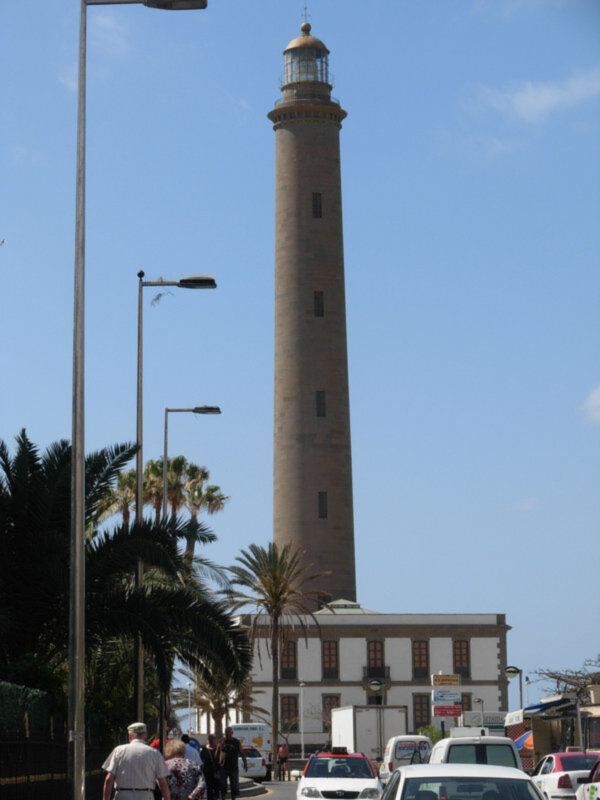 Lighthouse in the background with palm trees and people walking in the foreground.