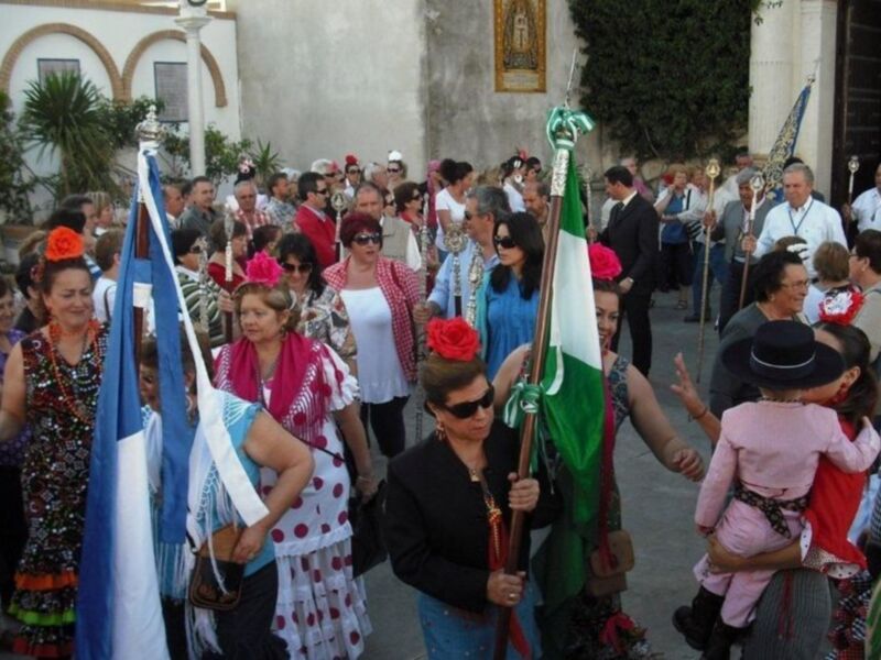 Fiesta tradicional con mujeres vestidas de trajes típicos andaluces, portando banderas y palmas, en un entorno urbano.