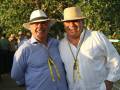 Dos hombres sonrientes con sombreros y joyas, posando juntos en un evento al aire libre.