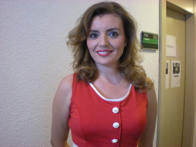 Una mujer con cabello ondulado y largo, vistiendo una camiseta roja con botones blancos, sonríe frente a un fondo de oficina.