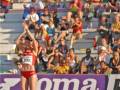 Una atleta con un top rojo y shorts verdes levanta la mano en señal de victoria mientras una multitud observa desde las gradas. La imagen se centra en el momento de la celebración, con una bandera de Roma visible al fondo.