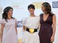 Tres mujeres sonrientes, con dos de ellas sosteniendo un premio, en un evento publicitario con anuncios de bebidas y productos de lujo en el fondo.