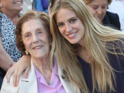 Una anciana sonríe junto a una joven con cabello largo y rubio. Ambas parecen estar en un evento formal, posando para una foto junto a otras personas en el fondo.
