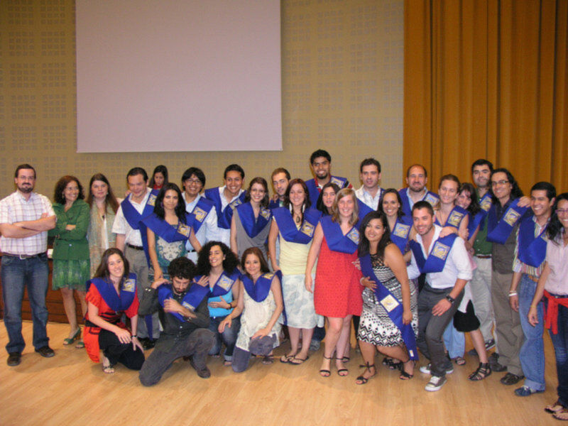 Grupo de personas posando juntos en un evento formal, todos con chalecos azules y vestidos de gala. En el fondo, se puede ver una pantalla blanca y cortinas amarillas. La imagen transmite una sensación de unidad y celebración.