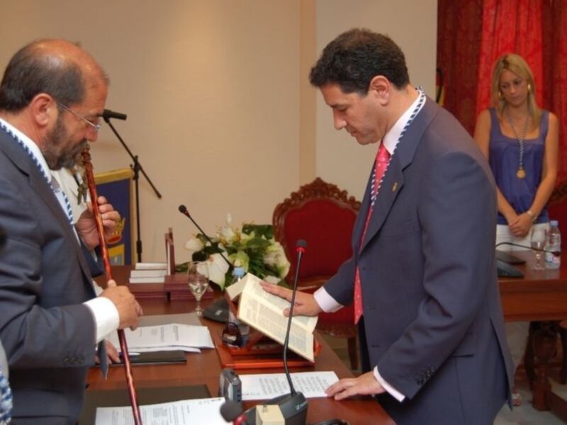 Dos hombres en trajes se encuentran en una sala formal, uno leyendo un libro mientras el otro observa. La sala está decorada con una bandera y flores, y hay una mujer en el fondo. El ambiente sugiere una reunión o conferencia formal.