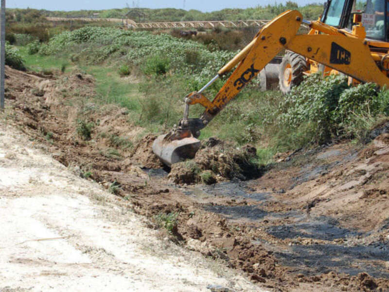 Excavadora amarilla trabajando en un canal de drenaje, con tierra y agua alrededor.
