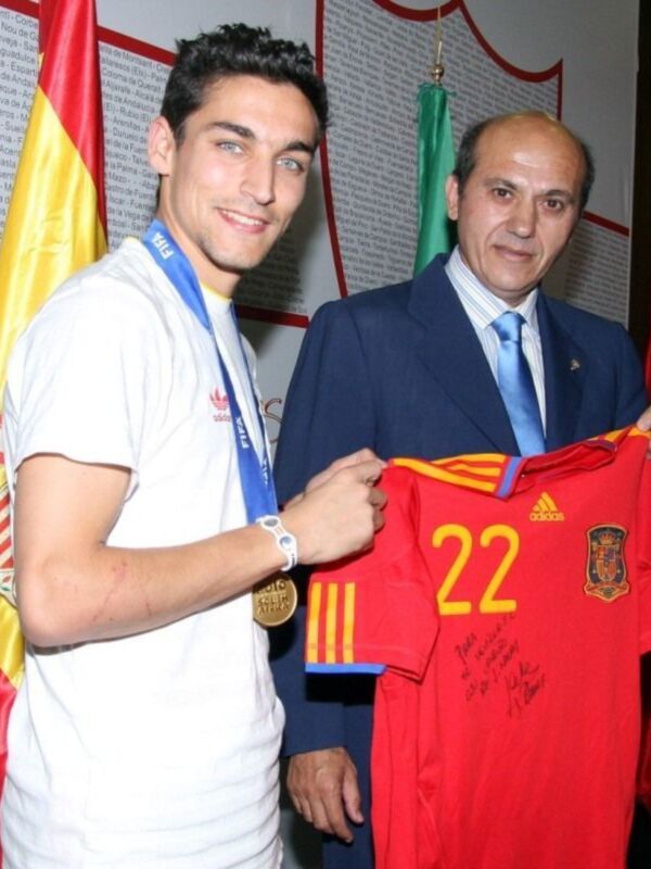 Un joven sostiene una camiseta de la selección española con el número 22, mientras un hombre en traje sostiene una bandera española. Ambos están frente a una pared con texto y banderas de diferentes países.