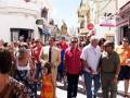 Una multitud camina por una calle con tiendas y edificios de colores vibrantes. Algunos llevan sombreros, pantalones cortos y camisetas de colores. La escena parece ser una celebración o feria local, con gente disfrutando del ambiente.