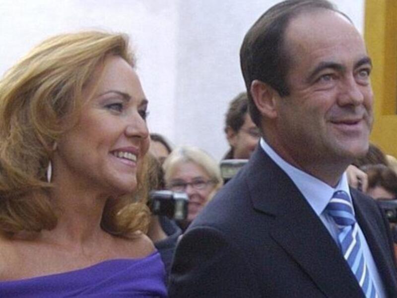 Dos personas sonrientes, una mujer con cabello largo y una persona masculina en traje. La imagen parece ser de un evento público, con personas a su alrededor y una camara fotográfica en primer plano.