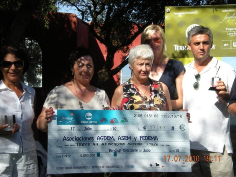Una foto de un grupo de personas sosteniendo un cheque grande, con una fecha y números en él, en un evento público con una pancarta en el fondo.