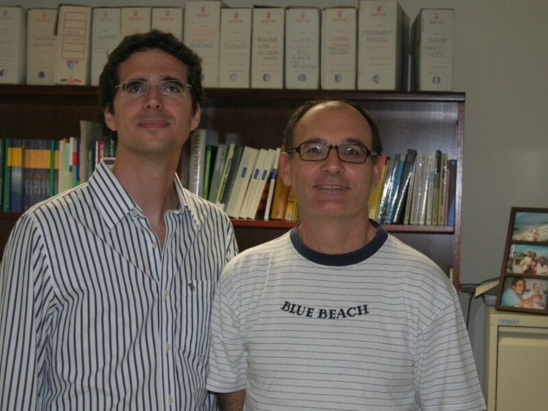 Dos hombres sonrientes frente a una estantería llena de libros. El hombre en el centro lleva un jersey con la palabra "BLUE BEACH" y sonríe ampliamente.