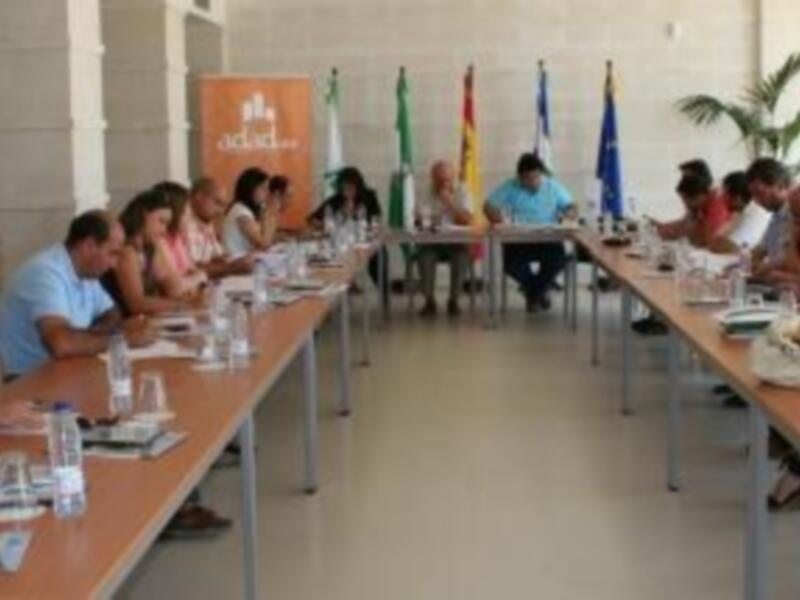 Una reunión formal en una sala de conferencias con varias personas sentadas a mesas, sosteniendo documentos y charlando. La sala tiene una decoración clásica con un póster de la Universidad de Alcalá en el fondo.