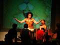 Una actriz en un vestido rojo se encuentra en el centro de la escena, con un fondo proyectado que muestra una imagen de un personaje antiguo. La escena parece ser parte de una representación teatral o una actuación.