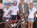 Un ciclista con una camiseta blanca y verde, un hombre en traje y dos mujeres sonrientes posan junto a una bicicleta roja con detalles blancos. El fondo muestra un póster de Andalucía y una estructura blanca con la palabra "Andalucía" repetida.