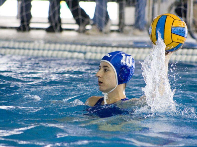 Un niño en un traje de baño azul y gorro de natación azul está jugando al waterpolo. La pelota amarilla y azul de Adidas está en el aire, justo encima del agua. El niño parece estar lanzando la pelota hacia otro jugador que está fuera de la imagen.