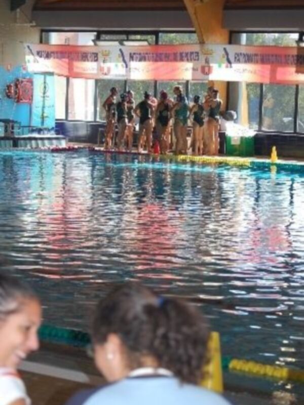 Una piscina con nadadores en la línea de salida, personas observando desde el borde y un marco de fotos con texto en la pared.