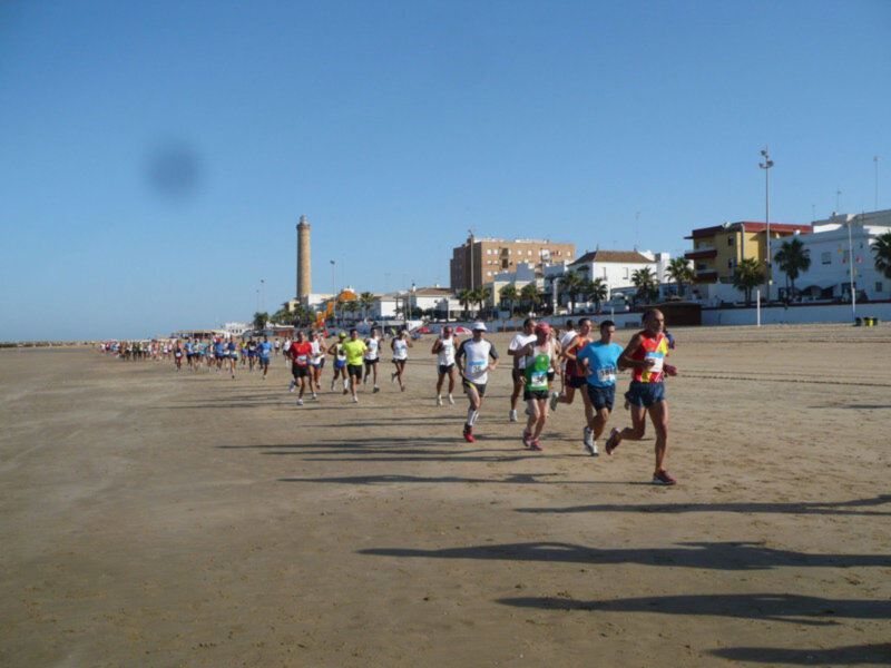 Maratón en la playa, con corredores en diferentes posiciones. En el fondo, edificios y una torre de vigilancia.
