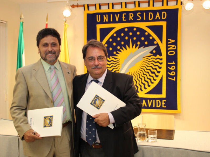 Dos hombres en trajes sostienen documentos en una sala con bandera de la Universidad AVIDE y el año 1997 en el fondo.