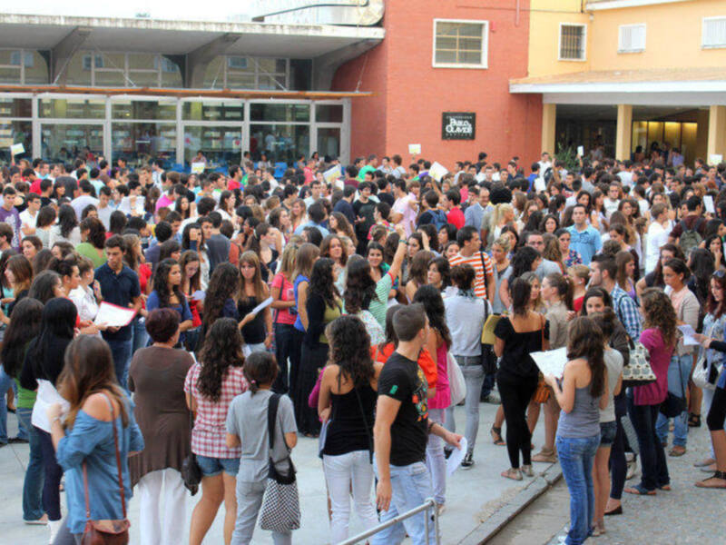 Un grupo de personas reunidas en una plaza, posiblemente en una ceremonia o evento público, con edificios de colores distintivos al fondo.