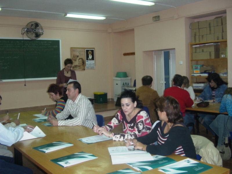Clase en una aula con estudiantes sentados en mesas, un profesor frente al pizarra y estantes con libros.