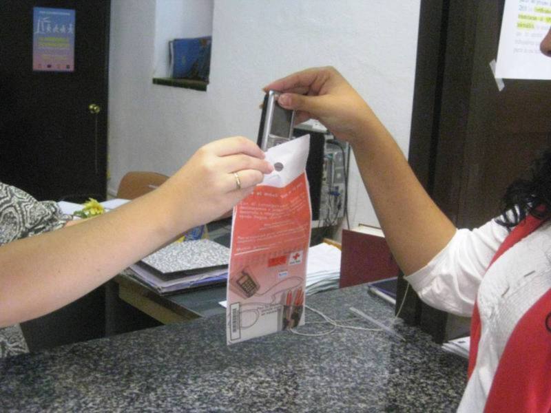 Una persona sostiene un teléfono móvil en una mano mientras otra persona le muestra un paquete de productos. La escena se desarrolla en un entorno de oficina con una computadora y otros elementos de escritorio visibles.