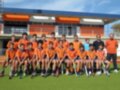 Un equipo de hockey infantil en uniforme naranja posa frente a una cancha deportiva. El equipo está sentado en fila, con los jugadores sosteniendo palos de hockey. Al fondo, se puede ver una estructura deportiva con una cubierta azul y naranja.