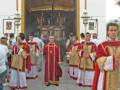 Un grupo de sacerdotes vestidos con trajes ceremoniales rojos y dorados procesiona por una calle, seguidos de fieles. La imagen muestra detalles de la vestimenta y el ambiente festivo, con un fondo que parece ser una iglesia o templo.