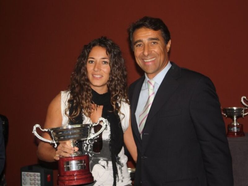 Una mujer y un hombre sonríen mientras sostienen trofeos. La mujer lleva una camisa blanca con detalles de blanco y negro, mientras que el hombre está vestido formalmente. El fondo es rojo oscuro con una mesa de trofeos en el lado derecho.