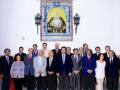 Una reunión formal de varios hombres y mujeres, todos vestidos con trajes formales, posan frente a una imagen religiosa de la Virgen María. La escena se centra en el respeto y la celebración de una ceremonia importante.