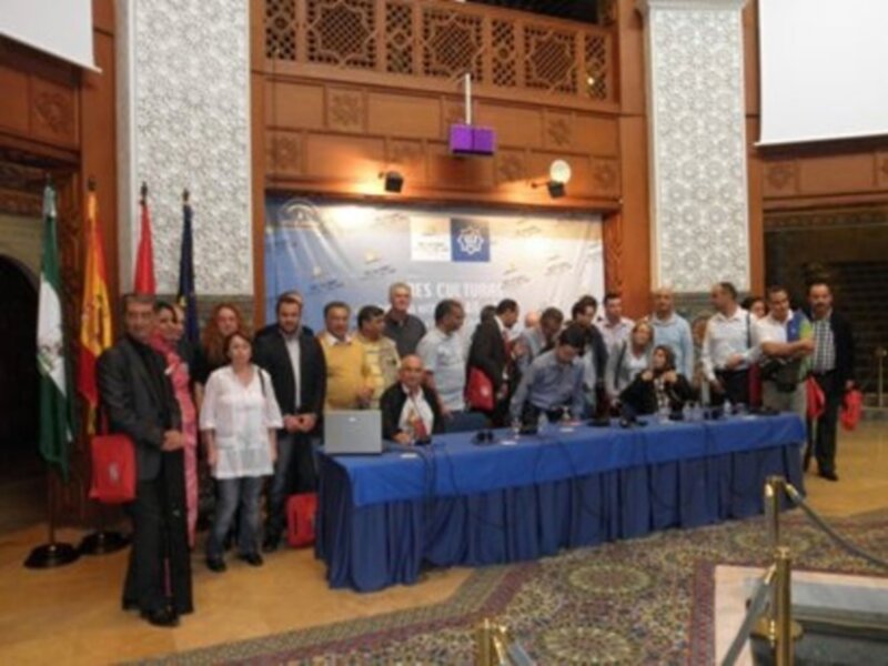 Una conferencia de prensa en un edificio con banderas y una mesa con micros y portátiles. Los asistentes están listos para la transmisión de noticias.