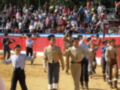 Un grupo de niños vestidos con trajes formales y sombreros se dirigen hacia la cámara, mientras un adulto toma una foto. El fondo muestra una multitud de espectadores en la tribuna, con un escenario rojo y blanco.