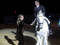 Una mujer y un hombre montan juntos en un caballo blanco, ambos vestidos formalmente. La escena parece ser nocturna con luces de fondo y un poste luminoso en el fondo.