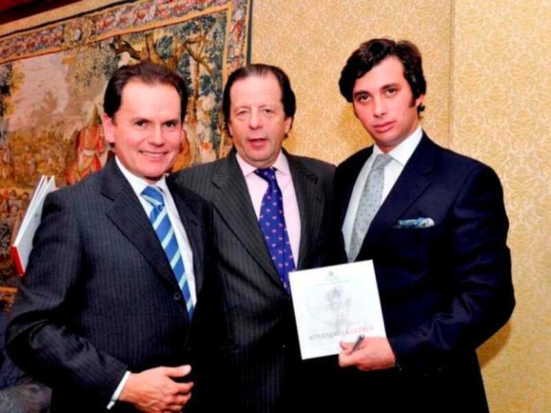 Tres hombres en trajes posan juntos, uno sostiene un libro. El fondo muestra una pintura de paisaje y papel tapiz.