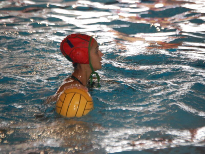Una persona jugando waterpolo en un estanque con una pelota amarilla y un casco rojo.