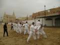 Un grupo de personas en ropa blanca practican artes marciales en una playa arenosa. En el fondo, se puede ver una iglesia con un campanario y edificios amarillos.