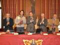 Una ceremonia de premiación en un salón con cortinas rojas y doradas. Un hombre sostiene un trofeo mientras otros tres hombres aplauden. La mesa es roja con un emblema dorado y el fondo muestra una imagen de un personaje histórico.