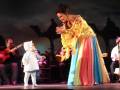 Una mujer vestida con un traje tradicional colorido se encuentra en escena, mientras un niño pequeño y una persona adulta tocan instrumentos musicales.